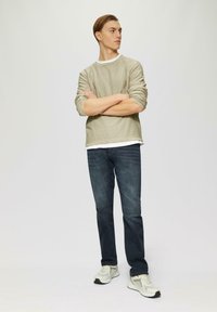 Lichtbeige langemouwen shirt laag over een wit T-shirt, gecombineerd met donkerblauwe jeans en witte sneakers met grijze accenten.