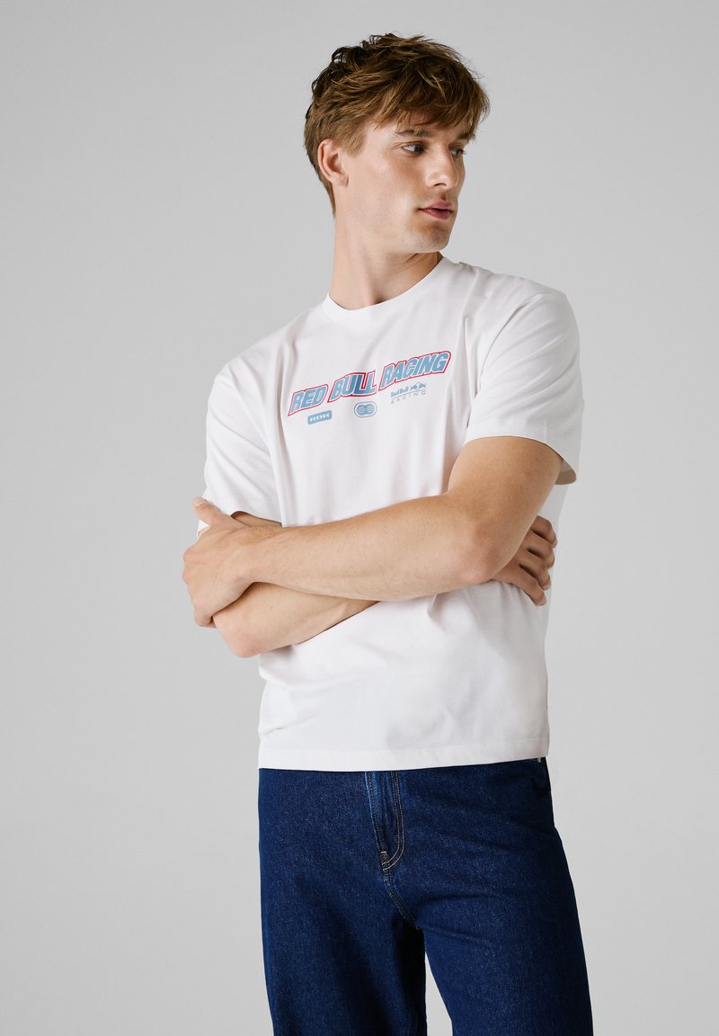 Biała bawełniana koszulka z graficznym nadrukiem w czerwonym i niebieskim, na którym widnieje napis "RED BULL RACING", zestawiona z ciemnoniebieskimi dżinsami.