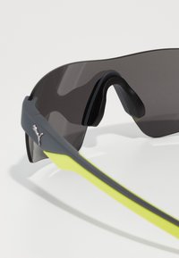 Lunettes de sport avec une monture noire mate et des accents jaunes vifs. Elles possèdent des verres teintés foncés et un design aérodynamique et élégant.