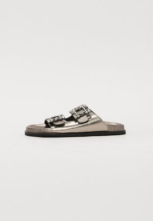 Sandalias deslizantes metálicas plateadas con dos hebillas adornadas, diseño abierto en la parte delantera y una suela texturizada negra. Superficie lisa con acentos mínimos.