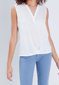White sleeveless blouse with a V-neck, featuring a subtle floral pattern and gathered design at the front. Paired with light blue pants.