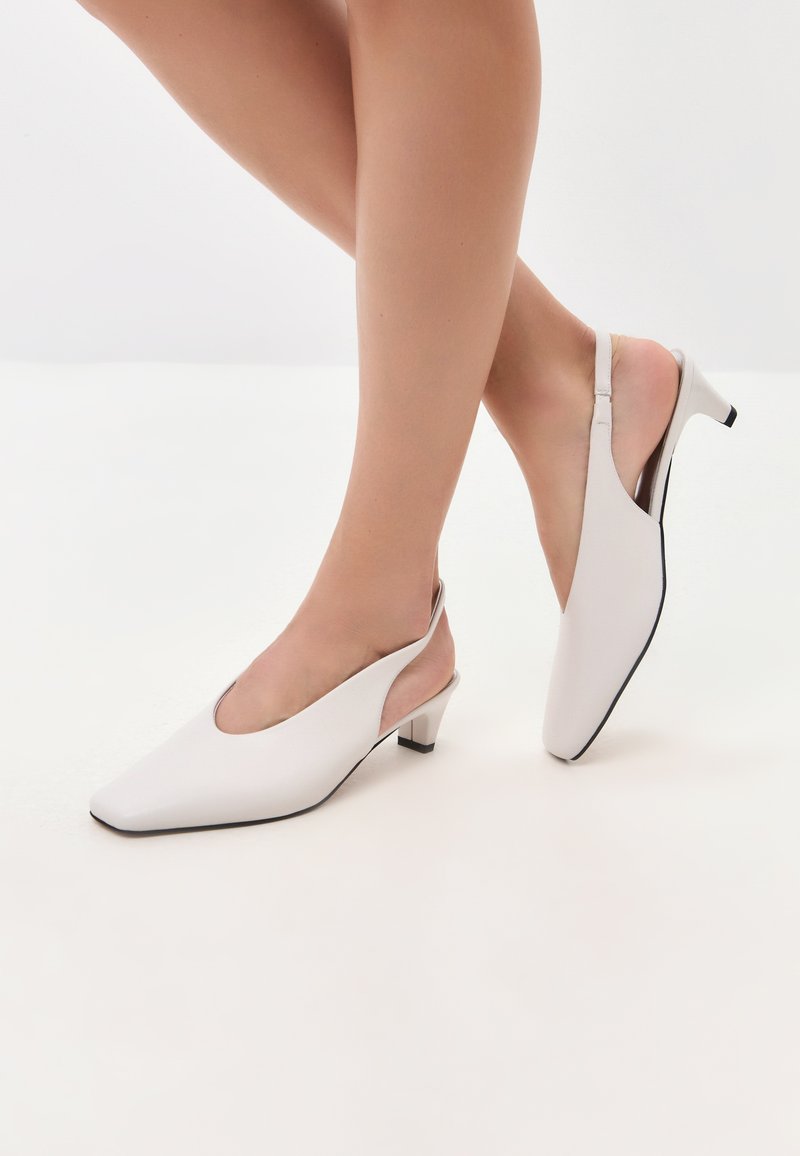 Pantofi slingback albi cu bot pătrat, toc mic și laturi deschise. Textură netedă cu accente de margine negre.