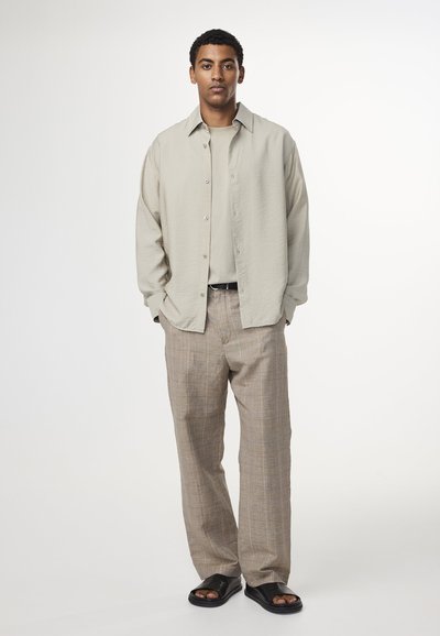 Ljus beige skjorta med knappar över en ljus taupe T-shirt, tillsammans med rutig bruna byxor och svarta flip-flop sandaler. Släta tygtexturer.