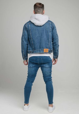 Homme debout dos à la caméra portant une veste en denim bleue par-dessus un sweat à capuche gris, un jean bleu et des baskets blanches sur fond uni.
