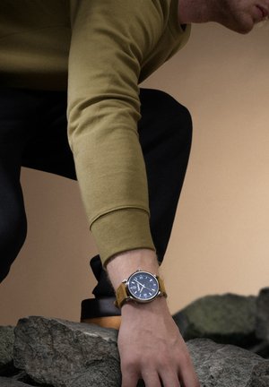 Orologio rotondo in argento con quadrante blu, numeri bianchi e cinturino in pelle gialla senape, indossato su una mano che si allunga verso delle rocce.
