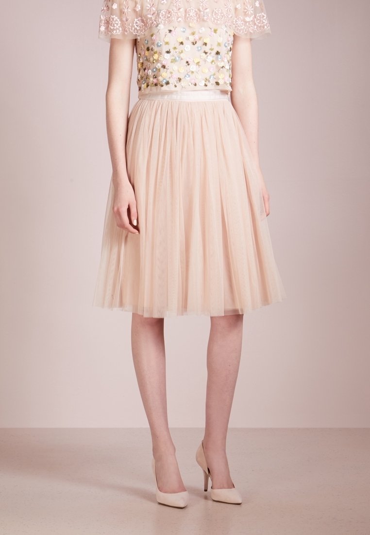Femme portant une jupe midi en tulle rose pâle, un haut orné de fleurs avec manches en dentelle, et des chaussures à talons hauts rose pâle assorties.