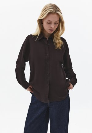 Blondynka ubrana w czarną koszulę na guziki z przednią kieszenią oraz luźne ciemnoniebieskie dżinsy, patrząca w dół z rękami w kieszeniach.