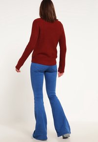 Maglione rosso a coste con maniche lunghe abbinato a jeans a zampa d'elefante blu, caratterizzato da una texture liscia e una vita aderente. Vista dalla schiena.
