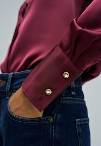 Blusa de satén burdeos con acentos de botones dorados en los puños, que presenta una textura suave y un diseño ajustado, combinada con jeans azul oscuro.