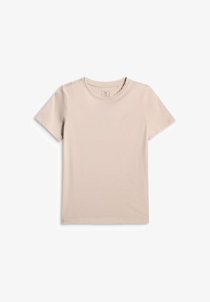 Jeune fille portant une chemise beige, un pantalon de survêtement gris et un sweat-shirt posé sur les épaules, tenant un sac bandoulière beige sur fond gris.