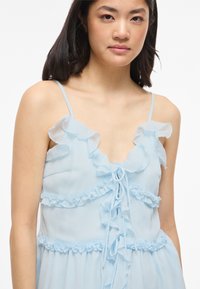 Jeune femme portant une robe sans manches bleu clair avec des bords volantés et fines bretelles sur fond blanc.
