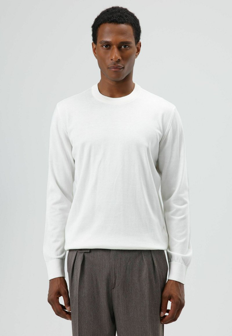 Wit shirt met lange mouwen gemaakt van zacht materiaal, met een ronde hals en geribbelde manchetten, gecombineerd met grijze geplooide broek, geposeerd tegen een neutrale achtergrond.