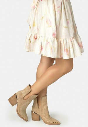 Ben gekruist met beige suède enkellaarzen met blokhakken en studs, gecombineerd met een lichte bloemenrok met ruches.