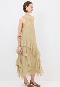 Beige Chiffonkleid mit geschichteten Rüschen, Halterneck-Ausschnitt und fließender Silhouette. Kombiniert mit olivgr�ünen spitzen Ballerinas.