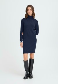 Marinblå mock-turtleneck klänning med långa ärmar, ribbad textur, slim fit och sidologodetalj. Kombinerad med knähöga svarta stövlar.
