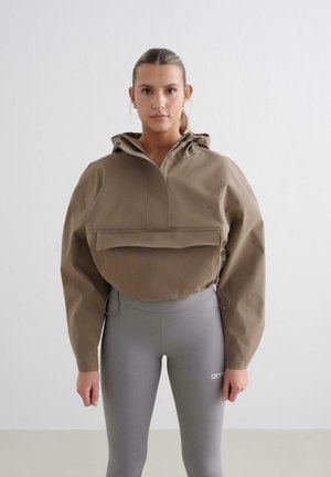 Ruda trumpa striukė su gaubtu, turinti priekinę kišenę ir elastines detales. Derinama su šviesiai pilkais legginsais, ant kurių matomas logotipas.
