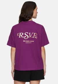 Vrouw met lang bruin haar, gekleed in een paarse T-shirt met gouden tekst "R.S.V.P fila tennis society" en zwarte shorts, met haar rug naar de camera.