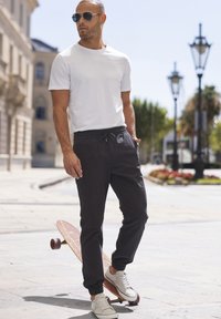 Witte T-shirt met korte mouwen, zwarte joggerbroeken met elastische boorden en witte sneakers. Skateboard met houten structuur bij de voeten op een verharde ondergrond.