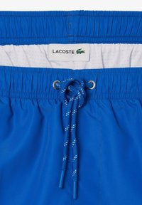 Bañador azul con cinturilla elástica, forro de malla blanca y cordón anudado. Etiquetado "LACOSTE" con el logo de un cocodrilo.