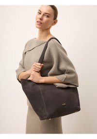 Szürke velúrbőr tote táska arany kiegészítőkkel, széles nyílással, puha tapintással és masszív vállpánttal. A modell a táskát egy világos háttér előtt tartja.