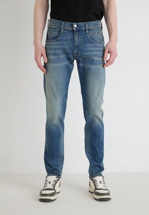 Mand iført falmede blå jeans og hvide sneakers står mod en ensfarvet lys baggrund med hænderne afslappet ned langs siderne.