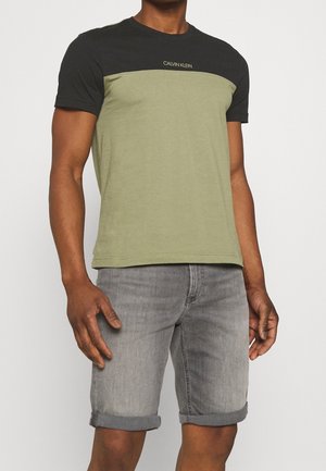 Mand iført en sort og olivengrøn Calvin Klein t-shirt med grå optrukne denimshorts mod en ensfarvet baggrund.