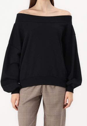 Femme portant un chemisier noir à manches longues épaules dénudées avec des manches volumineuses et un pantalon marron à carreaux taille haute.