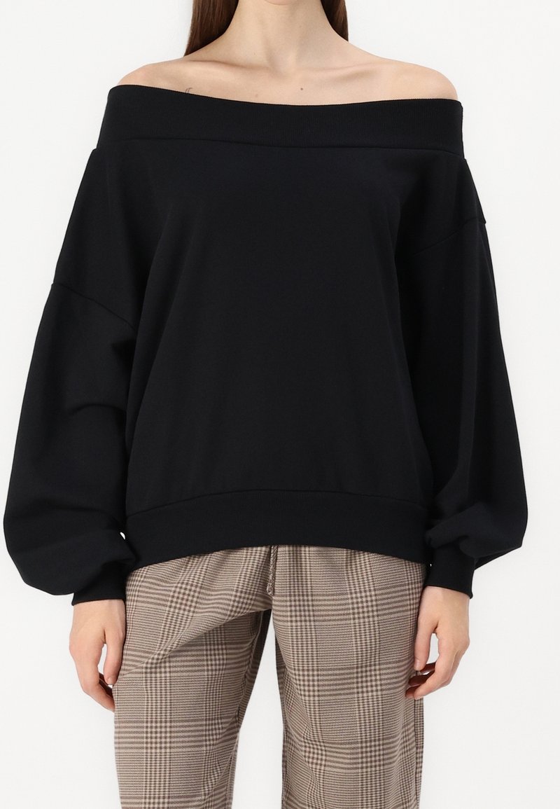 Femme portant un chemisier noir à manches longues épaules dénudées avec des manches volumineuses et un pantalon marron à carreaux taille haute.