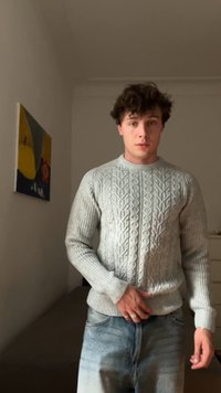 Ung mand med krøllet hår iført en grå kabelstrikket sweater og lyseblå jeans, der står indendørs nær en seng og vægkunst.