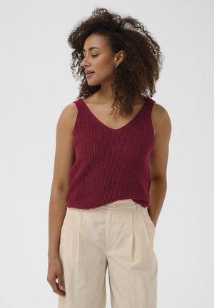 Femme aux cheveux bouclés portant un haut en maille bordeaux sans manches et un pantalon beige taille haute, debout avec une main dans la poche.