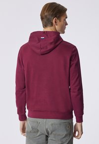 Felpa cappuccio color bordeaux con una consistenza morbida. Presenta polsini e orlo a costine, con un piccolo logo in blu, bianco e rosso sulla schiena.