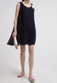 Yhden olkapään kukka-aksentilla varustettu laivastonsininen pleatattu hihaton mekko, yhdistettynä clutch-laukkuun ja vaaleanpunaisiin sandaaleihin, joissa on väljä, virtaava muotoilu.