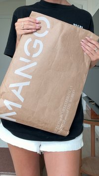 Brown paper bag with white text branding "MANGO," featuring a matte texture and slightly crumpled surface, held in a person's hands.
