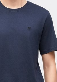 Marineblå bomulls t-skjorte med korte ermer, med en liten svart geometrisk logo på venstre brystområde. Jevn tekstur og klassisk snitt.