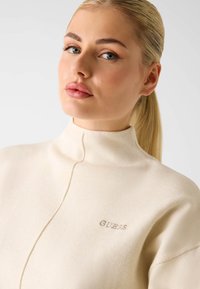 Samtig-cremefarbener Pullover mit hohem Kragen und einem dezenten, eingestickten "GUESS"-Logo. Glatte Textur, lockere Passform und klare Linien.