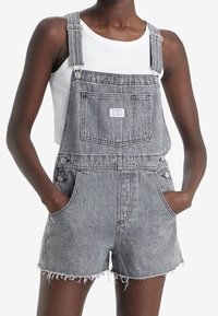 Graue Jeans-Overalls mit verstellbaren Trägern, seitlichen Knöpfen, einer Fronttasche und fransigen Säumen. Getragen über einem weißen Tanktop.