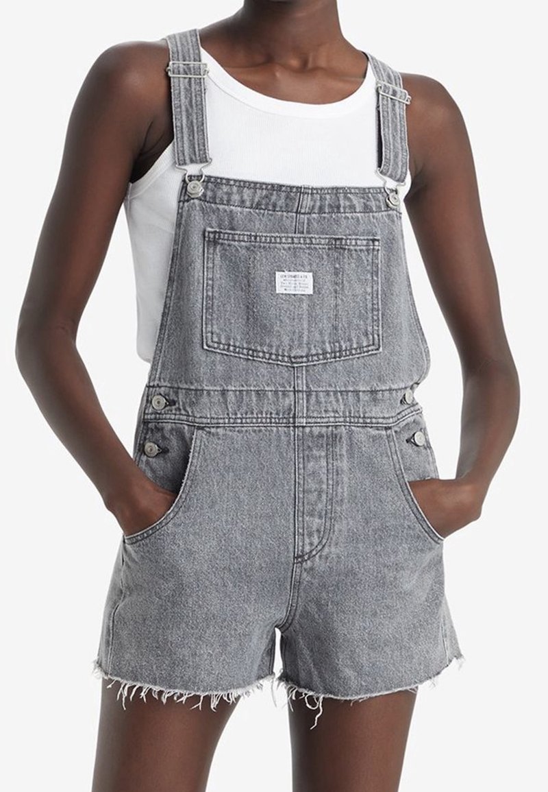 Graue Jeans-Overalls mit verstellbaren Trägern, seitlichen Knöpfen, einer Fronttasche und fransigen Säumen. Getragen über einem weißen Tanktop.