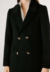 Cappotto doppiopetto in misto lana verde scuro con revers a punta, tasche laterali e bottoni dorati; tessuto texture con una finitura liscia.
