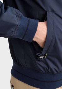 Námořnická modrá bunda s elastickými manžetami, kapsou na zip a zeleným akcentem. Hladký povrch s jemným kostkovaným vzorem a kontrastními pruhy.