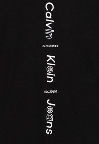 Černé tričko s vertikálním bílým textem: "Calvin Klein Established 40.730610" a "Jeans" se různě velkými písmovými velikostmi, čistým designem a měkkou texturou.