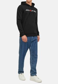 Schwarzer Kapuzenpullover mit weißem und rotem "JACK & JONES"-Logo, vordertasche in Känguru-Form, gerippte Bündchen, kombiniert mit blauen Jeans und weißen Turnschuhen.