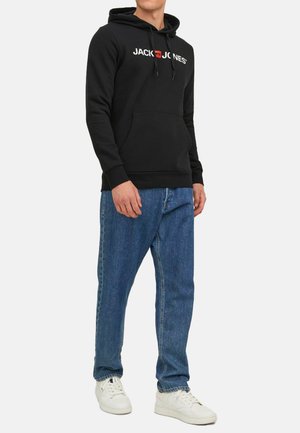 Schwarzer Kapuzenpullover mit weißem und rotem "JACK & JONES"-Logo, vordertasche in Känguru-Form, gerippte Bündchen, kombiniert mit blauen Jeans und weißen Turnschuhen.