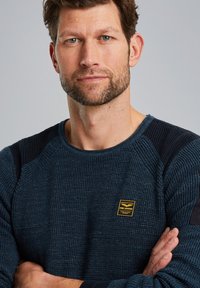 Dunkelblaues Sweatshirt mit Textur, das ein geripptes Muster, kontrastierende Schulterakzente und ein kleines gelbes Markenpatch auf der Brust aufweist.