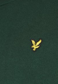 Maglione a coste di colore verde scuro con un piccolo logo di un uccellino giallo ricamato sul lato sinistro. La texture è morbida.