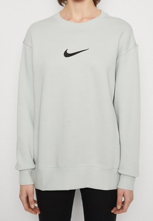 Person iført en lysegrå sweatshirt med lange ærmer med sort Nike Swoosh-logo centreret på brystet, kombineret med sorte bukser.