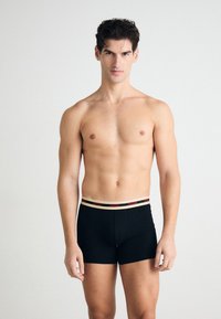 Boxers noirs en tissu extensible avec une ceinture beige arborant le logo rouge "HUGO". Le modèle se tient avec les bras détendus.
