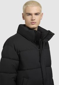 Schwarze Daunenjacke mit hohem Kragen, Reißverschluss und Seitentaschen; hat eine gestepptes Muster und ausgeprägte Polsterung.