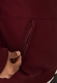 Bordeaux hoodie met een gladde textuur, voorzien van een ritszak aan de voorkant. Geribbelde boorden en onderrand accentueren het ontwerp.