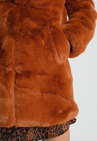 Pomarańczowy płaszcz z futerka syntetycznego o miękkiej fakturze, z zapięciem na guziki i bocznymi kieszeniami, prezentowany na jednolitym tle.