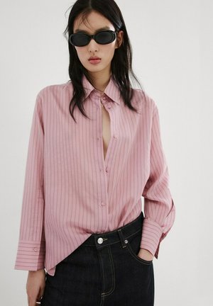 Mujer con camisa de botones rosa de rayas anchas, metida dentro de vaqueros oscuros, gafas de sol negras ovaladas, de pie con una mano en el bolsillo.